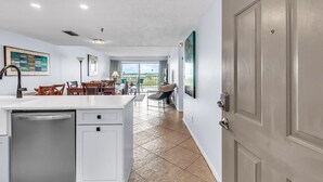 A modern kitchen with a dishwasher, a dining area with chairs and a table, and a view through a large window. - Sea Club #503 - Coastal Breeze (Indian Shores)
