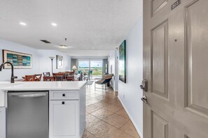 A modern kitchen with a dishwasher, a dining area with chairs and a table, and a view through a large window. - Sea Club #503 - Coastal Breeze (Indian Shores)