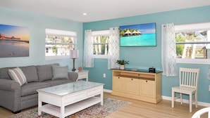 A living room with a sofa, a coffee table, a TV, and a cabinet. There are two windows with curtains, and a painting is on the wall. - Island Paradise Cottage (Fort Myers Beach)