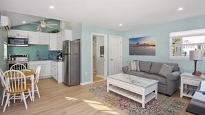 A modern kitchen with white cabinets, a stainless steel refrigerator, and a microwave. There's a dining area with a wooden table and white chairs. A gray sofa and a small white side table are in the living room, which has a large window and a beach scene painting. - Island Paradise Cottage (Fort Myers Beach)
