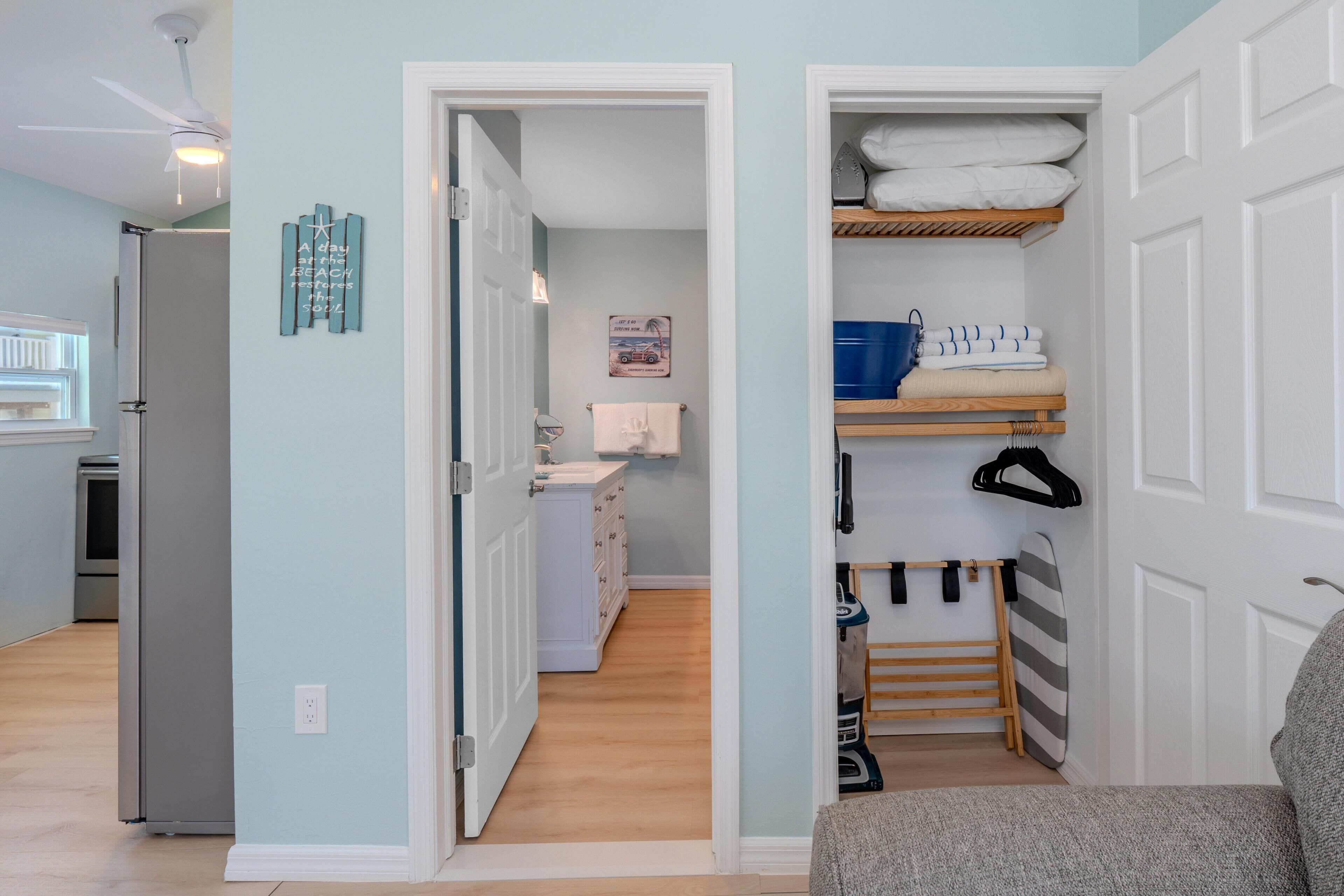 A room with a kitchenette, a bathroom with a towel rack, and a storage area with shelves and a vacuum cleaner.