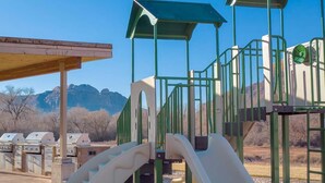 A playground with slides and climbing structures, barbecue grills, and a covered seating area with a view of mountains. - Spacious 2-bedroom condo with AC, WiFi in Magical Moab (Moab)