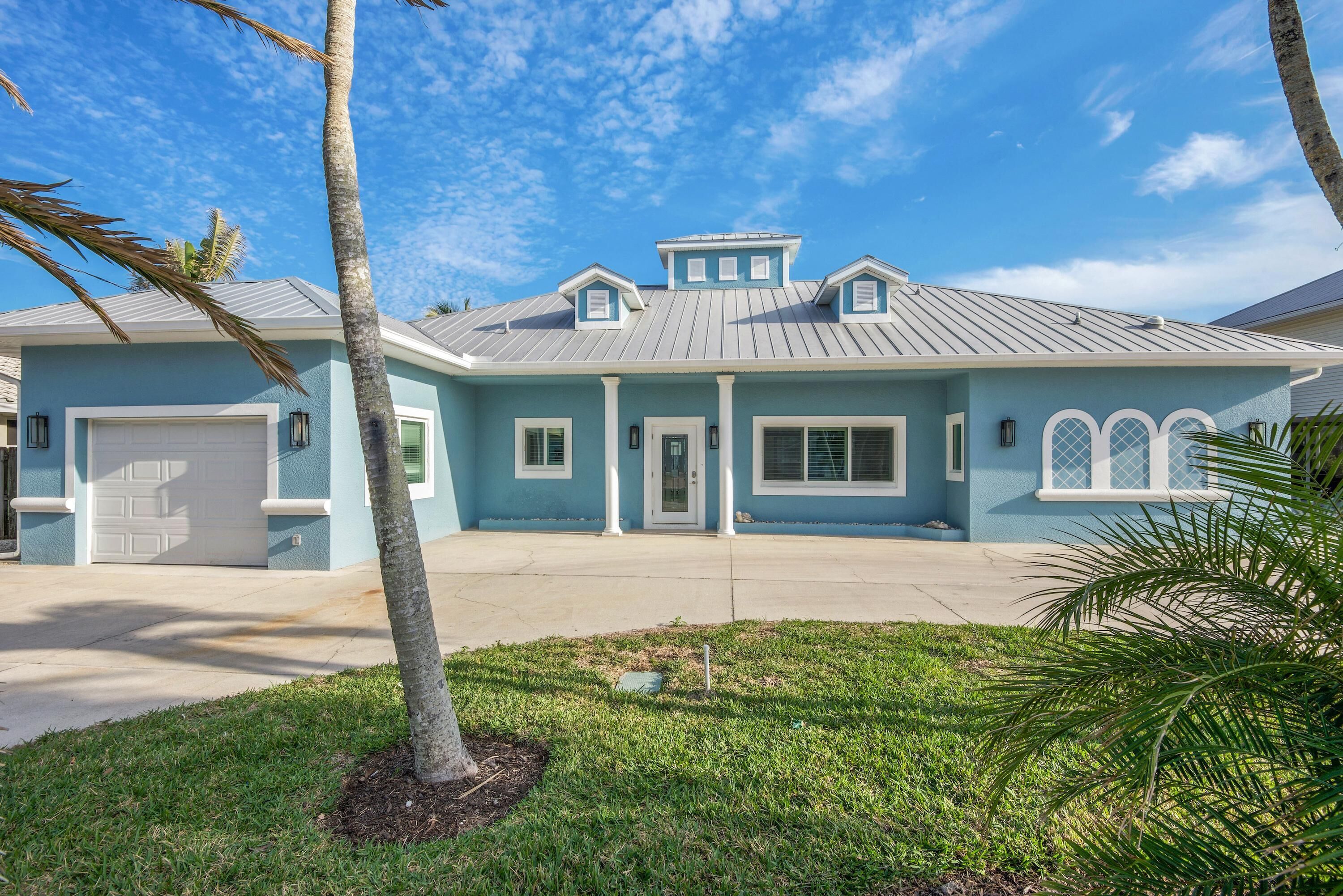 A two-story house with a light blue exterior, a garage, and a front yard with a palm tree.