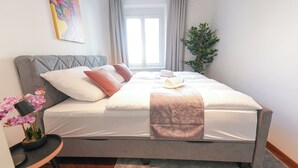 Una cama bien hecha con almohadas, una manta y una toalla enrollada. Hay una orquídea en maceta sobre una mesita de noche.