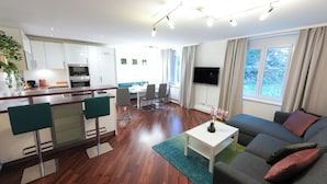 Una sala moderna con sofá, una mesa de centro y un comedor con sillas y una mesa. Hay una cocina con gabinetes blancos y un televisor montado en la pared.