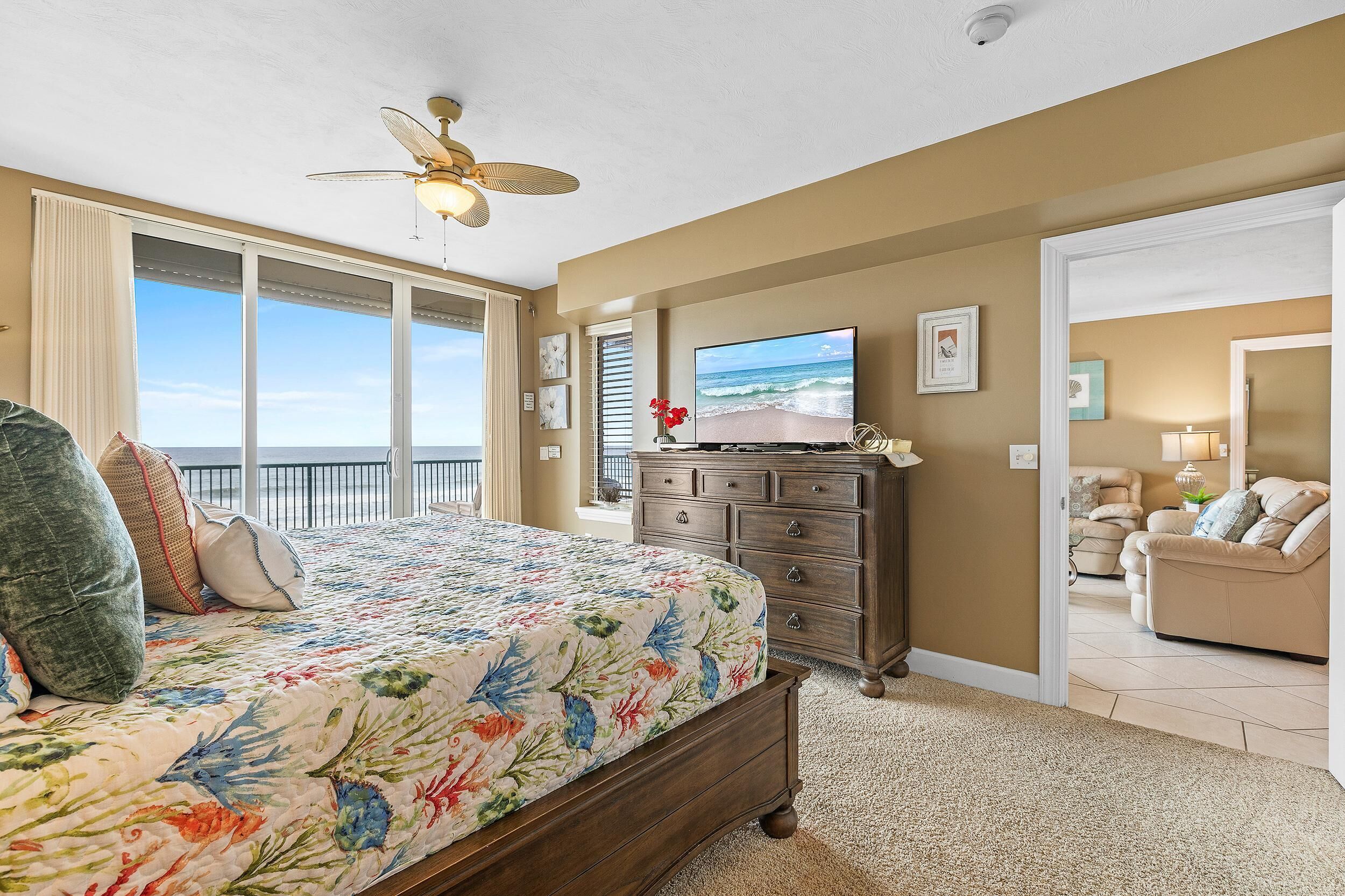 A bedroom with a large bed, a ceiling fan, a dresser, and a TV displaying a beach scene.