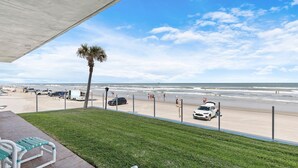 Blick auf einen Strand vom Balkon aus, mit einer Palme, einem weißen Auto und einem Promenadenweg am Strand.