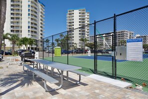 A tennis court with picnic tables and a fence, surrounded by tall buildings.
