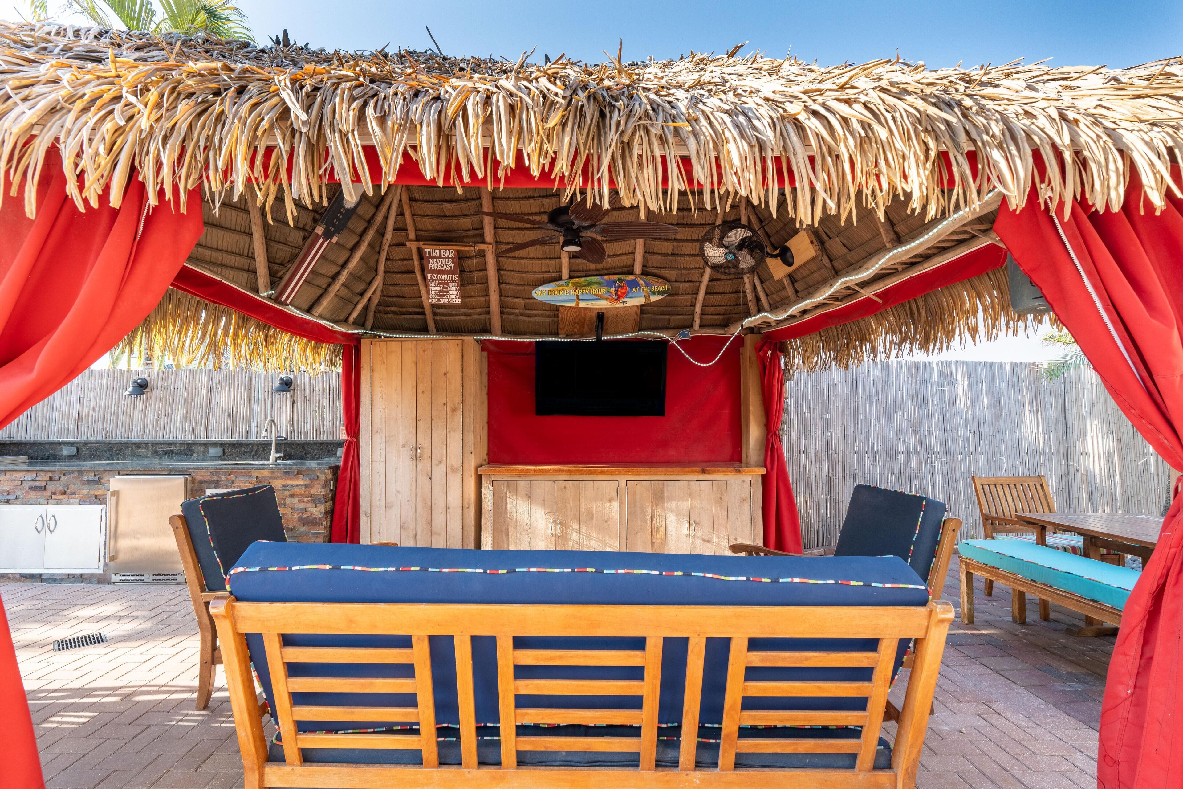 A thatched-roof outdoor seating area with wooden furniture and a red backdrop.