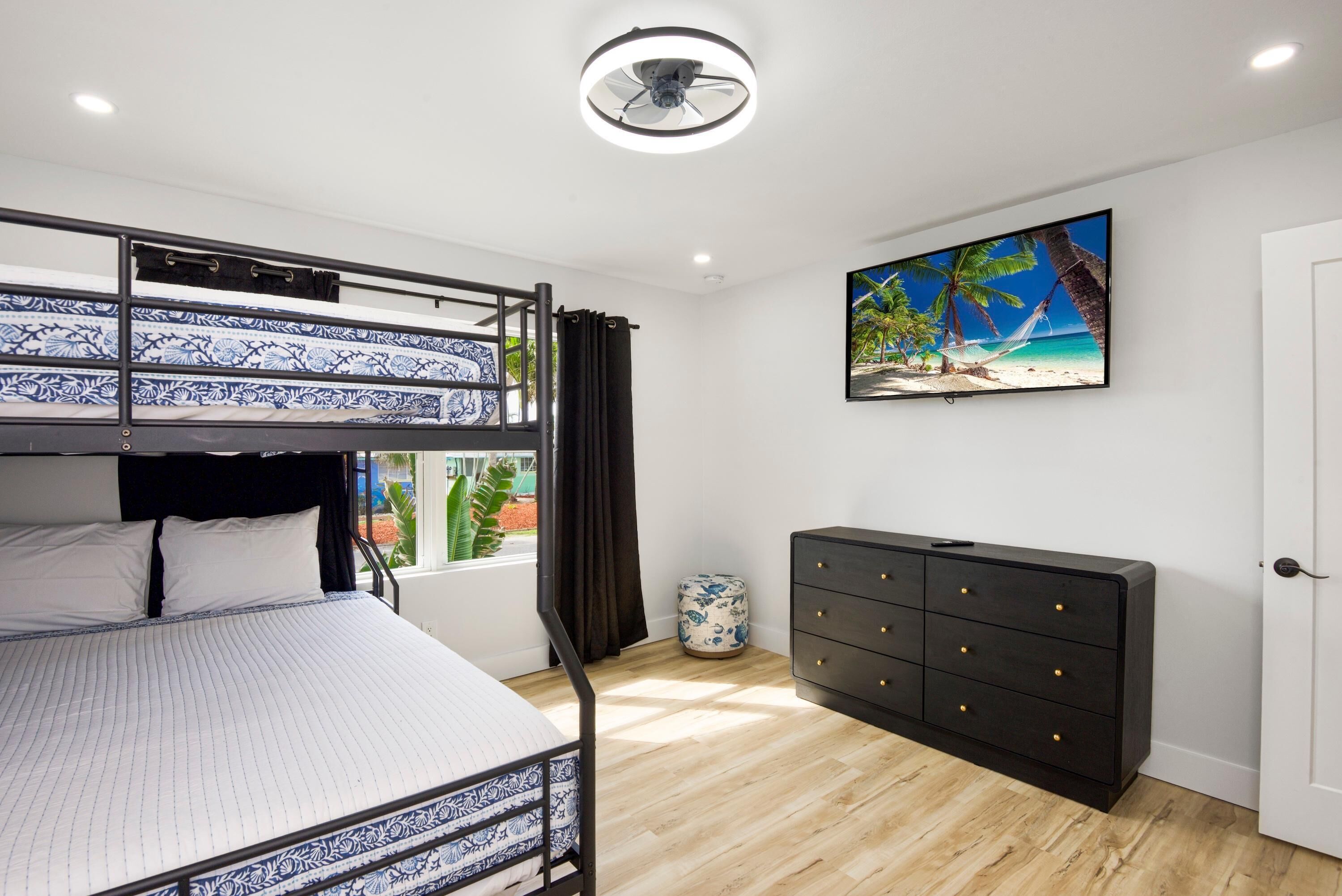 A room with a bunk bed, a dresser, a ceiling fan, a TV, and a window with curtains.