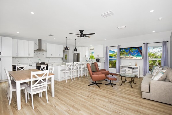 A modern kitchen with white cabinets, a dining area with a wooden table and white chairs, a living room with a sofa and a glass coffee table, and a large window with a beach scene.