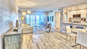 A spacious living area with a sofa, chairs, and a dining table. There is a kitchen area with a microwave and cabinets. The room has a large window and a view of the outdoors. - Land's End 1-206 Bay Front-premier (Treasure Island)