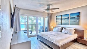 A modern hotel room with a large bed, a ceiling fan, a desk, and a balcony with a view of palm trees. - Land's End 1-206 Bay Front-premier (Treasure Island)