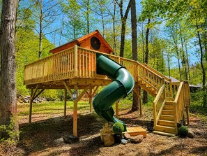 A wooden playhouse with a slide and stairs, surrounded by trees.