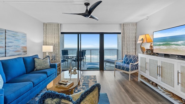 A modern living room with a blue sofa, a glass coffee table, and a television on a white cabinet. - Southwind 1002 (New Smyrna Beach)