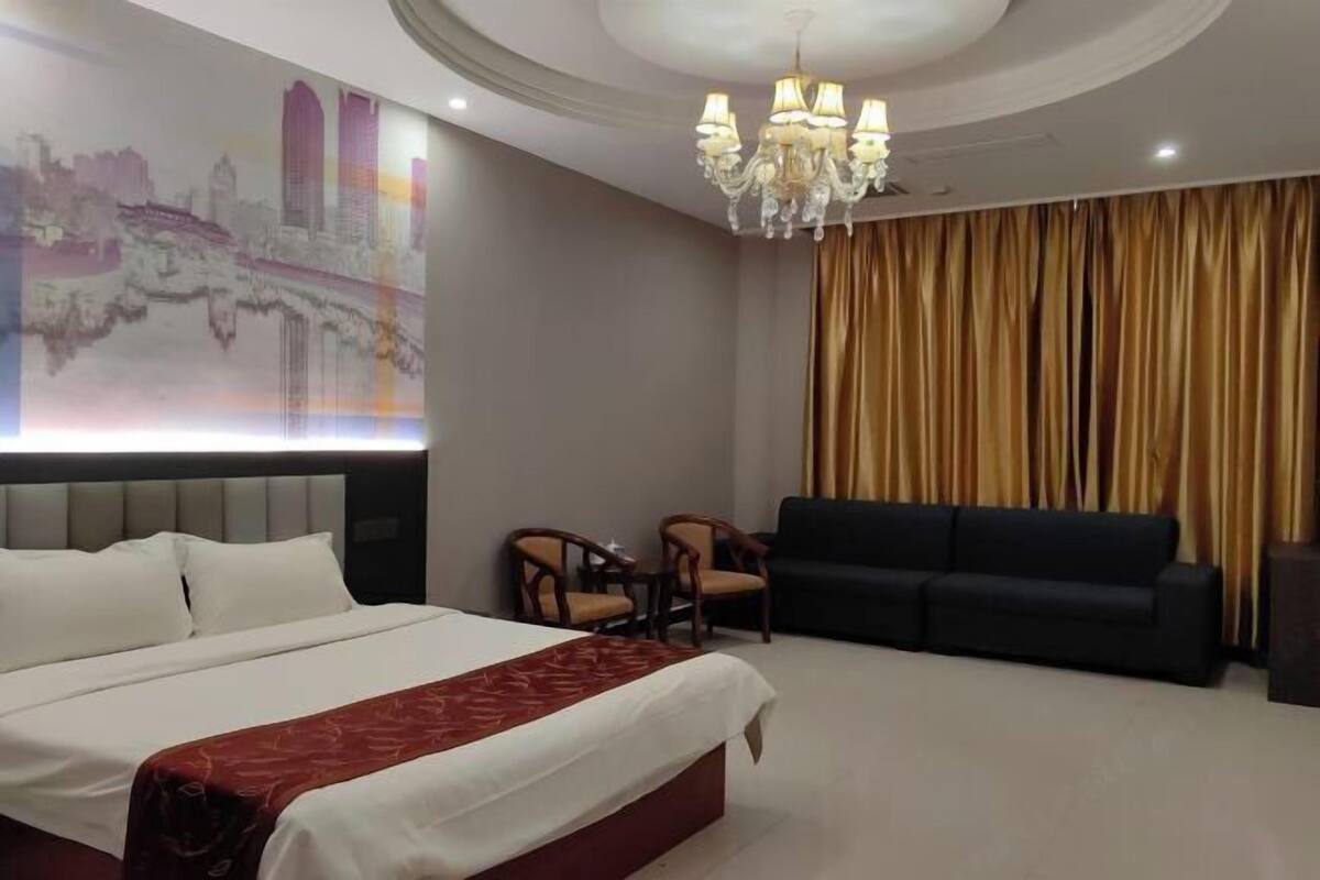 Habitación de hotel con una cama grande, un sofá, dos sillones, una araña y una pared con un mural de paisaje urbano.