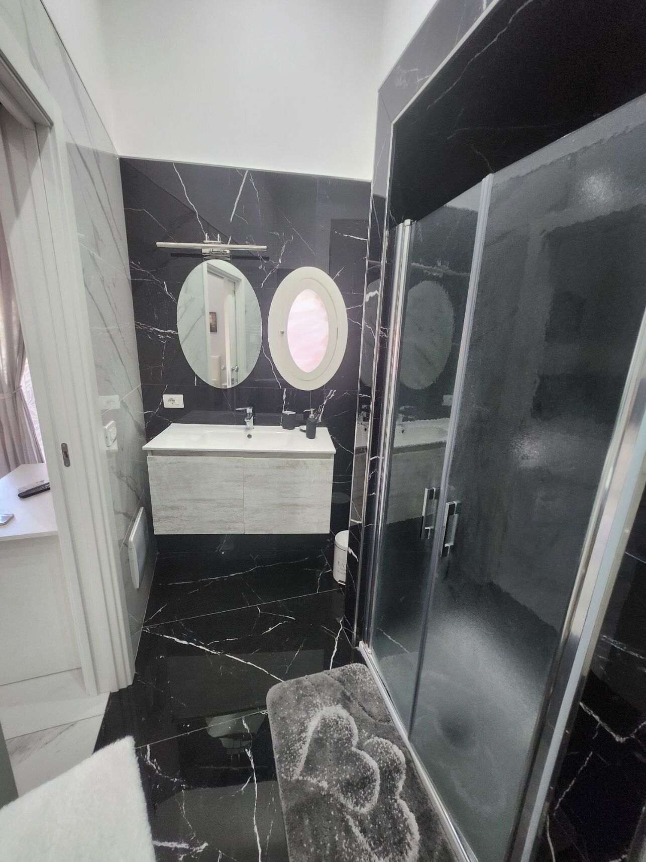 A modern bathroom with a black marble floor, a white vanity, two round mirrors, and a glass-enclosed shower area.