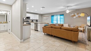 A modern kitchen with white cabinets, a central island, and a dining area with a brown sofa and a TV. - Oceans 25- Lazy Beach Days (Daytona Beach)