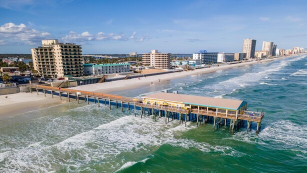 A pier extending into the ocean with a building on it, surrounded by a beach and city buildings. - Oceans 25- Lazy Beach Days (Daytona Beach)
