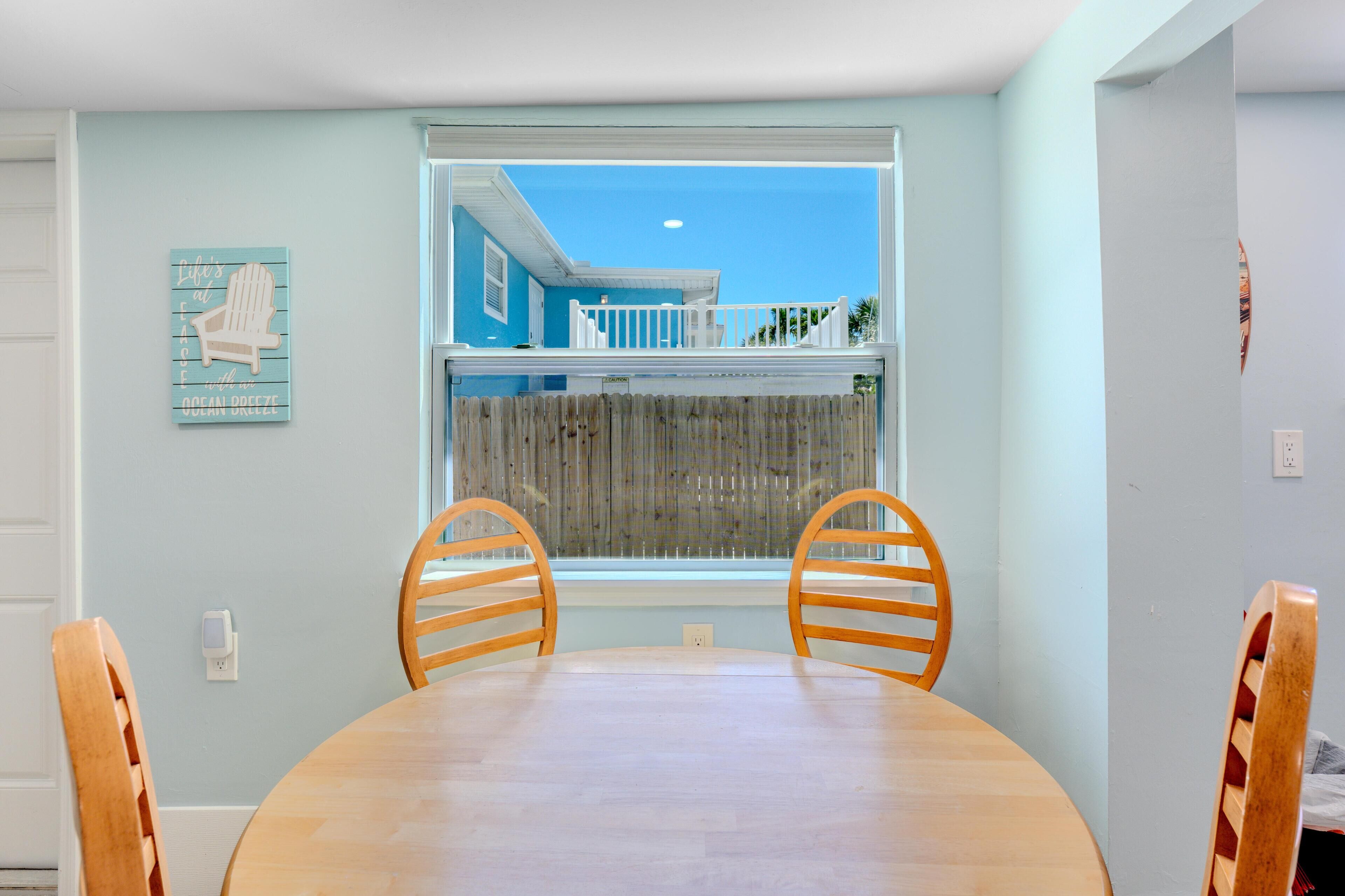 A dining area with a round wooden table and two wooden chairs, a window with a view of a house and a wooden fence, and a wall-mounted sign with text.