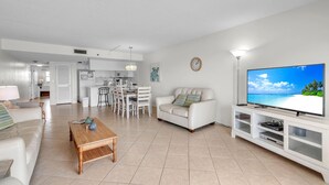 A modern living room with a white sofa, a wooden coffee table, a TV on a white stand, and a kitchen in the background. - Sea Club #402 - Isle Be Back (Indian Shores)