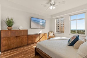 A bedroom with a wooden bed, a large window with a view of buildings, a ceiling fan, and a wall-mounted TV.