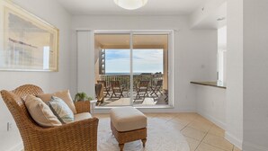 A room with a wicker chair, a small ottoman, and a view of an outdoor patio with a railing and chairs. - Minorca Paros 402 (New Smyrna Beach)