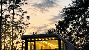 A modern, elevated cabin with a glass facade, two chairs, and a table, surrounded by trees at dusk. - Space Camp (Interlachen)
