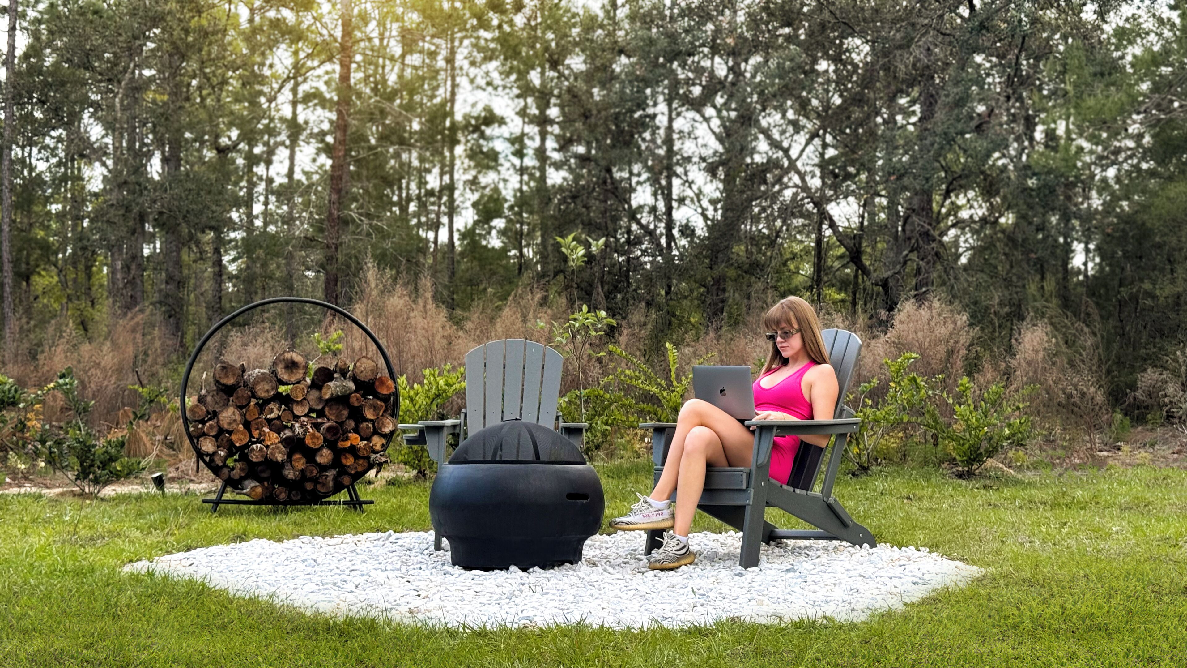 A person sitting on a chair in a grassy outdoor area with a laptop, surrounded by trees and a pile of logs.