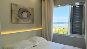 Une chambre à coucher avec un lit, une œuvre d’art accrochée au mur, une fenêtre avec des rideaux et une vue sur la plage.