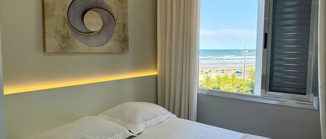 A bedroom with a bed, a wall-mounted artwork, a window with curtains, and a view of the beach.