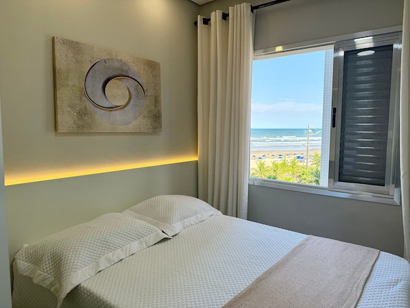 A bedroom with a bed, a wall-mounted artwork, a window with curtains, and a view of the beach.