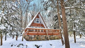A cabin with a stone foundation and a wooden deck, surrounded by snow-covered trees. - Cozy A-Frame ~ Ski Resorts • Game Room • EV • FirePit • Pets (pocono lake)