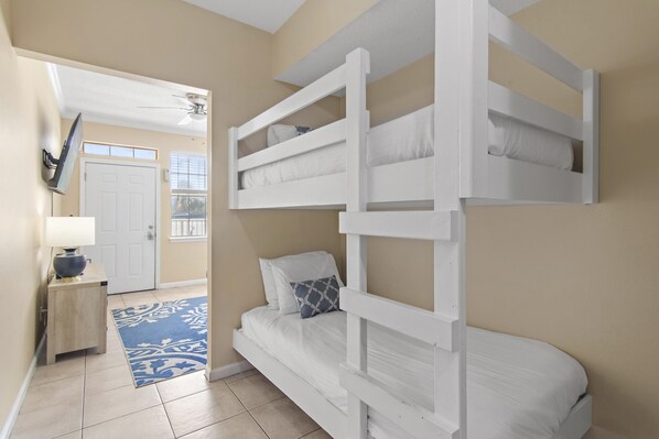 Une chambre avec un lit superposé, un tapis bleu à motifs, une petite table de chevet en bois avec une lampe, et une porte donnant sur une autre pièce.
