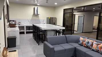 A modern kitchen with a marble countertop, a dining area with a white table and chairs, and a gray sofa with patterned cushions.
