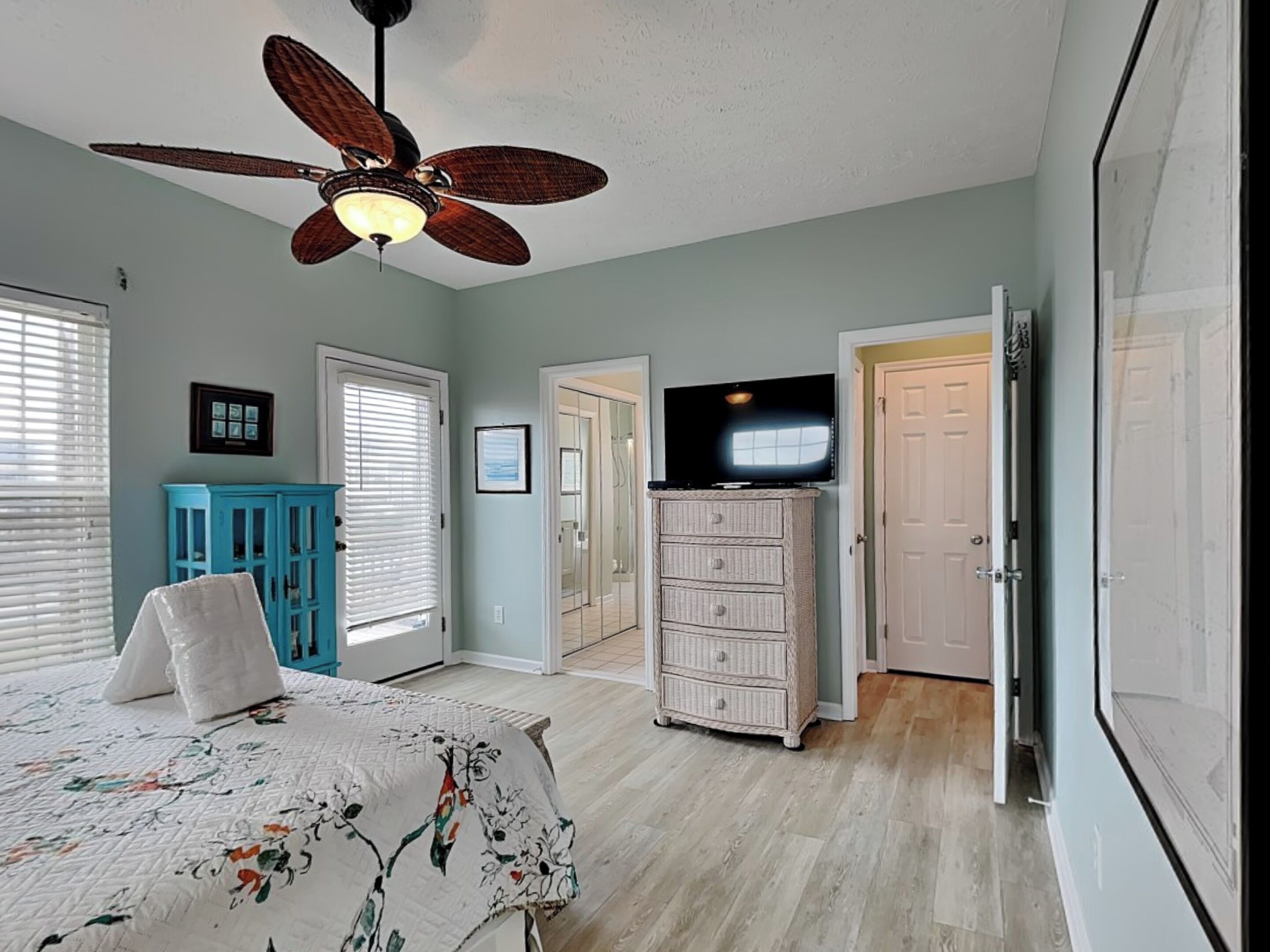 Une chambre à coucher comprenant un lit, un ventilateur de plafond, une commode, un téléviseur et une porte donnant sur une autre pièce.