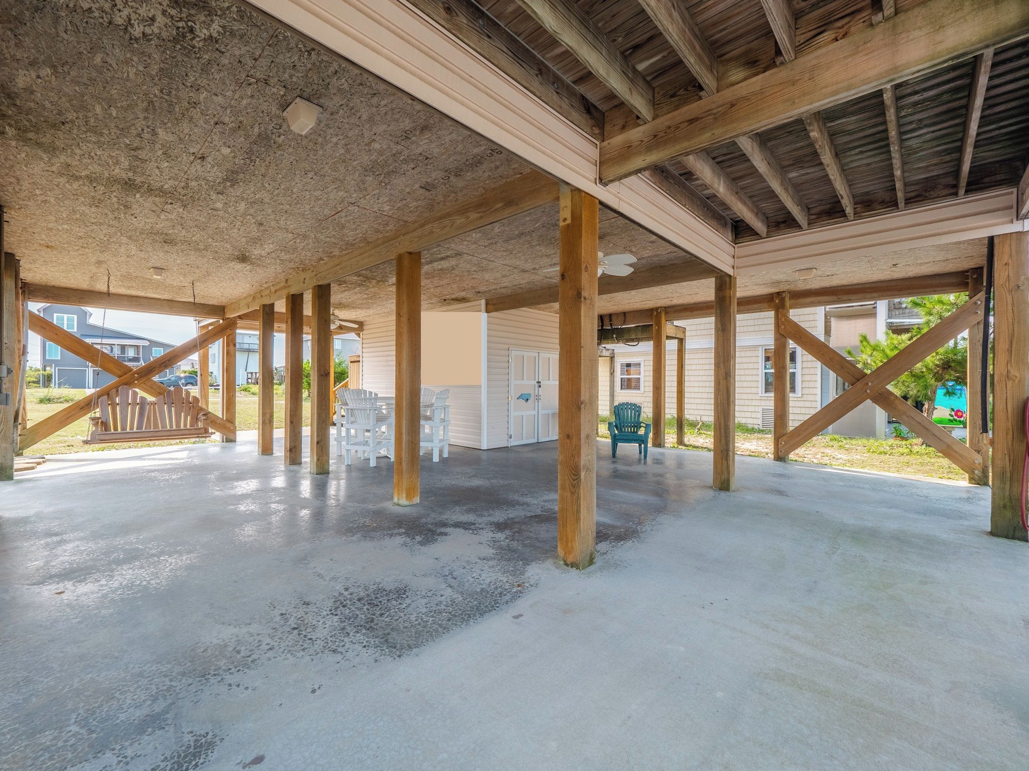 An open covered area with wooden beams and concrete flooring, featuring a swing set and outdoor seating.