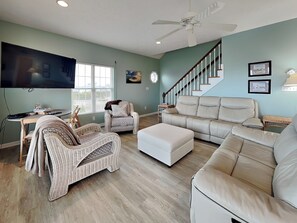 A spacious living room with a large flat-screen TV, wicker furniture, a beige leather sofa, and a staircase with a wooden railing. - Casa Capri by Topsail Realty (Holly Ridge)