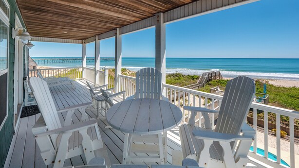 A covered outdoor seating area with white Adirondack chairs and a round table, overlooking a beach and a pier. - Shore Therapy by Topsail Realty (Sneads Ferry)