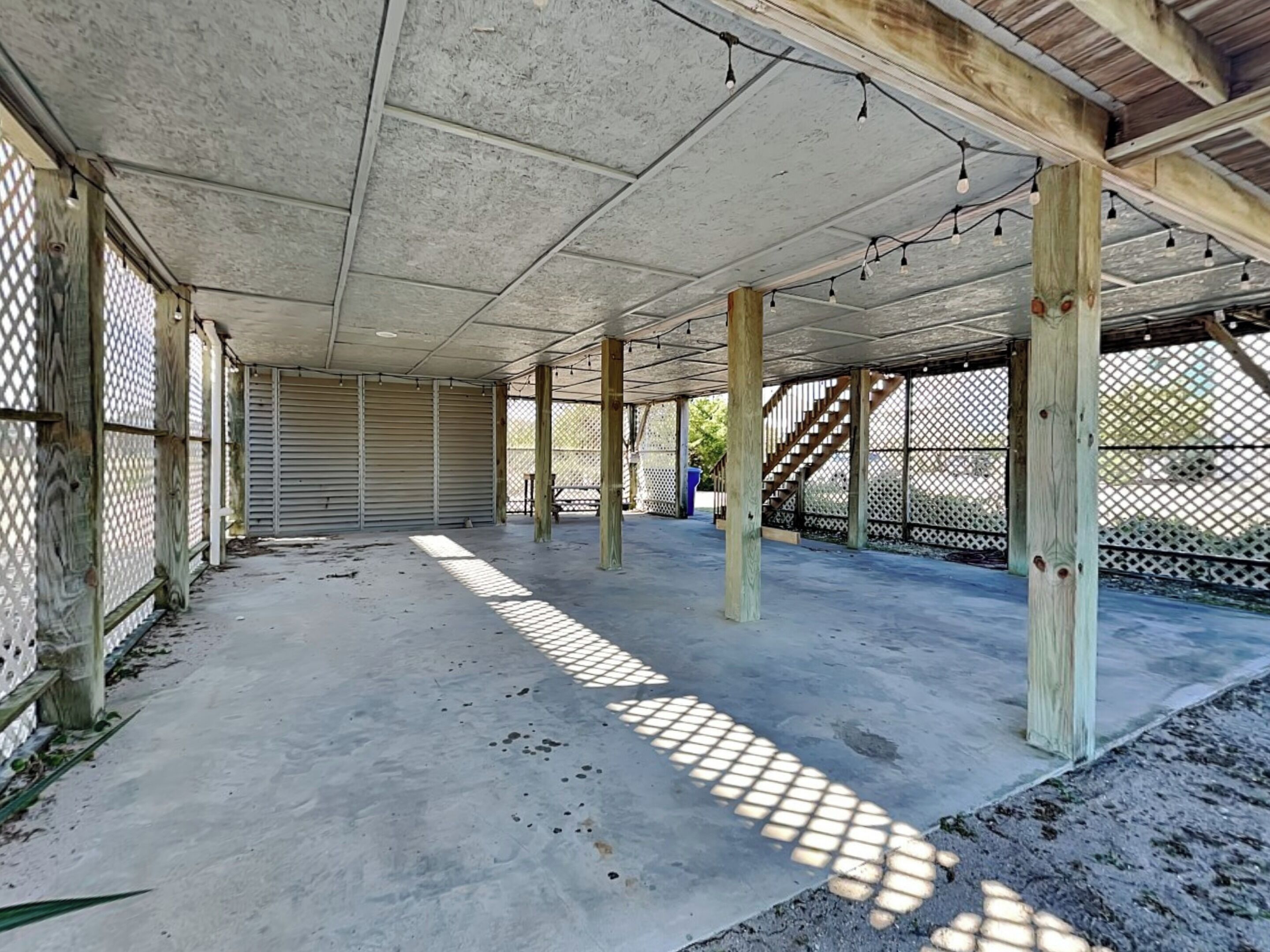 An open covered patio area with concrete flooring and wooden pillars, featuring a lattice fence and a view of the surrounding area.