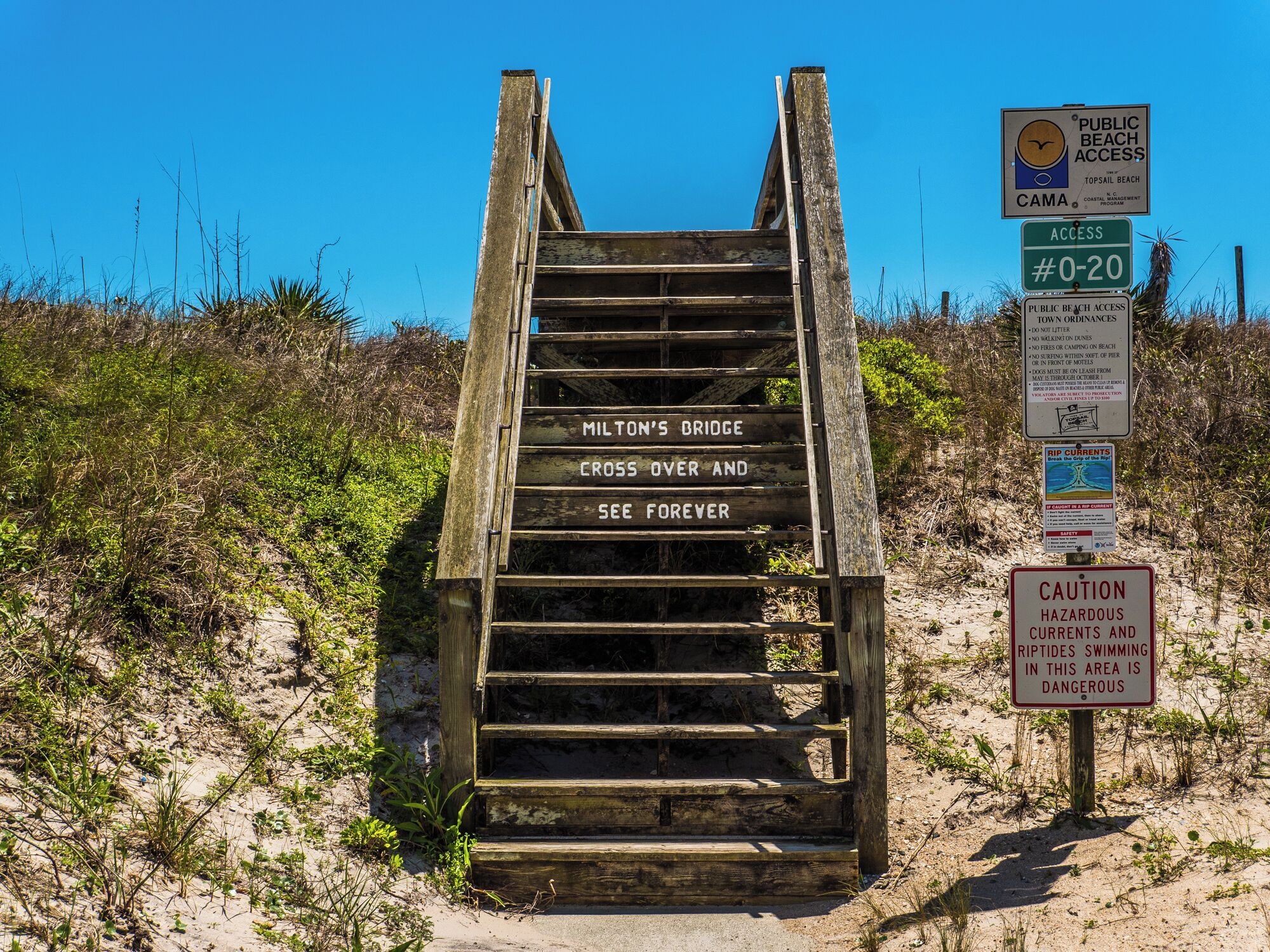 Una escalera de madera que conduce a una playa con varios carteles de advertencia e informativos.