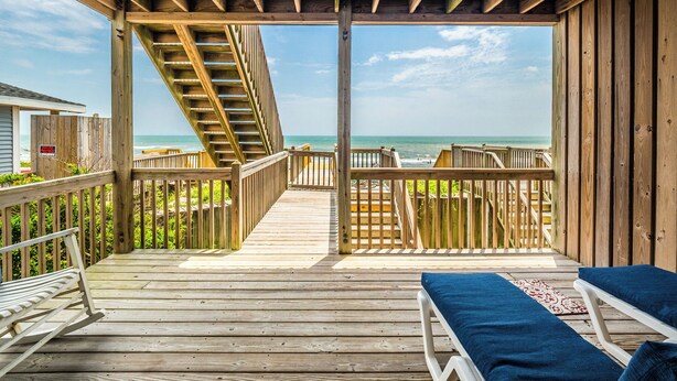 A wooden deck with a view of the ocean, a staircase, and two lounge chairs with blue cushions. - Hypnautical by Topsail Realty (Holly Ridge)