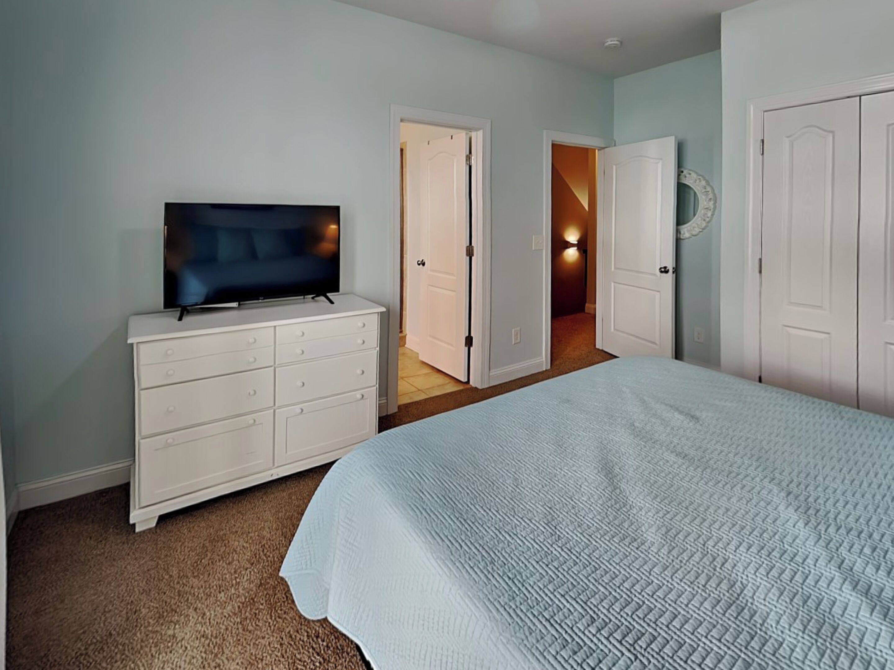 Quarto com cama, cômoda, televisão e duas portas que dão para outros espaços.