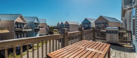 A wooden deck with a table and grill, overlooking a row of beach houses with balconies and a clear blue sky.