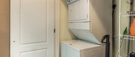 Une buanderie équipée d’un lave-linge et d’un sèche-linge superposés, d’une porte et d’une étagère contenant des produits de nettoyage.