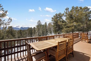 Eine Holzterrasse mit Tisch und Stühlen, mit Blick auf ein verschneites Bergmassiv und einen dichten Wald.