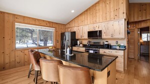 Una cocina moderna con armarios de madera, una isla central con taburetes de bar, electrodomésticos de acero inoxidable y un amplio ventanal con vistas a árboles.