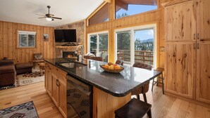 Interior de una cabaña de madera con cocina, comedor y un amplio ventanal con vistas a un paisaje montañoso.