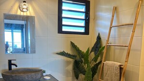A bathroom with a large mirror, a round sink, a window with blinds, a potted plant, a towel rack, and a waste bin.