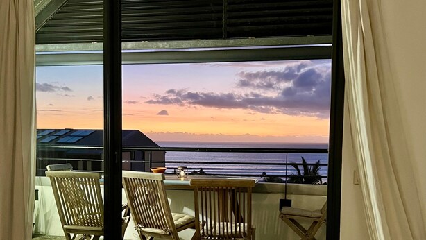 A room with a view of the ocean at sunset, featuring a table with chairs, a wooden bench, and a suitcase.
